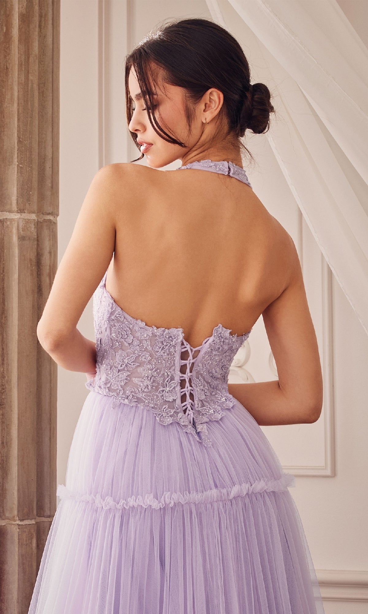 A woman stands with her back to the camera in the Lace-Halter Long A-Line Prom Dress A1206, highlighting its open back, lace-up detail, and delicate floral-lace bodice in a stunning lilac shade.